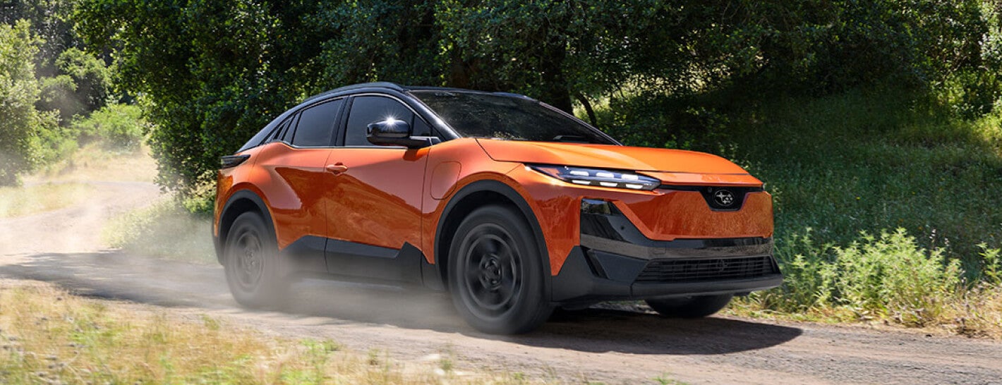 Front three-quarter view of an orange 2027 Subaru Uncharted EV driving on a dirt road surrounded by trees.