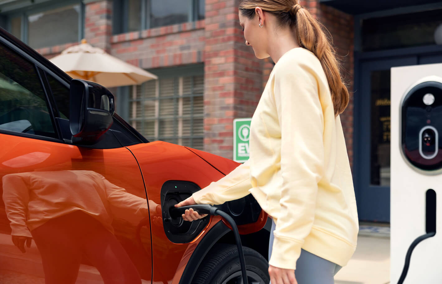 Close-up side view of an orange 2027 Subaru Uncharted EV at a charging station outside a brick building.