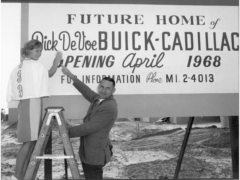 A man on a ladder and a woman stand next to a sign for the Future Home of Dick Devoe Buick-Cadillac opening in April 1968.