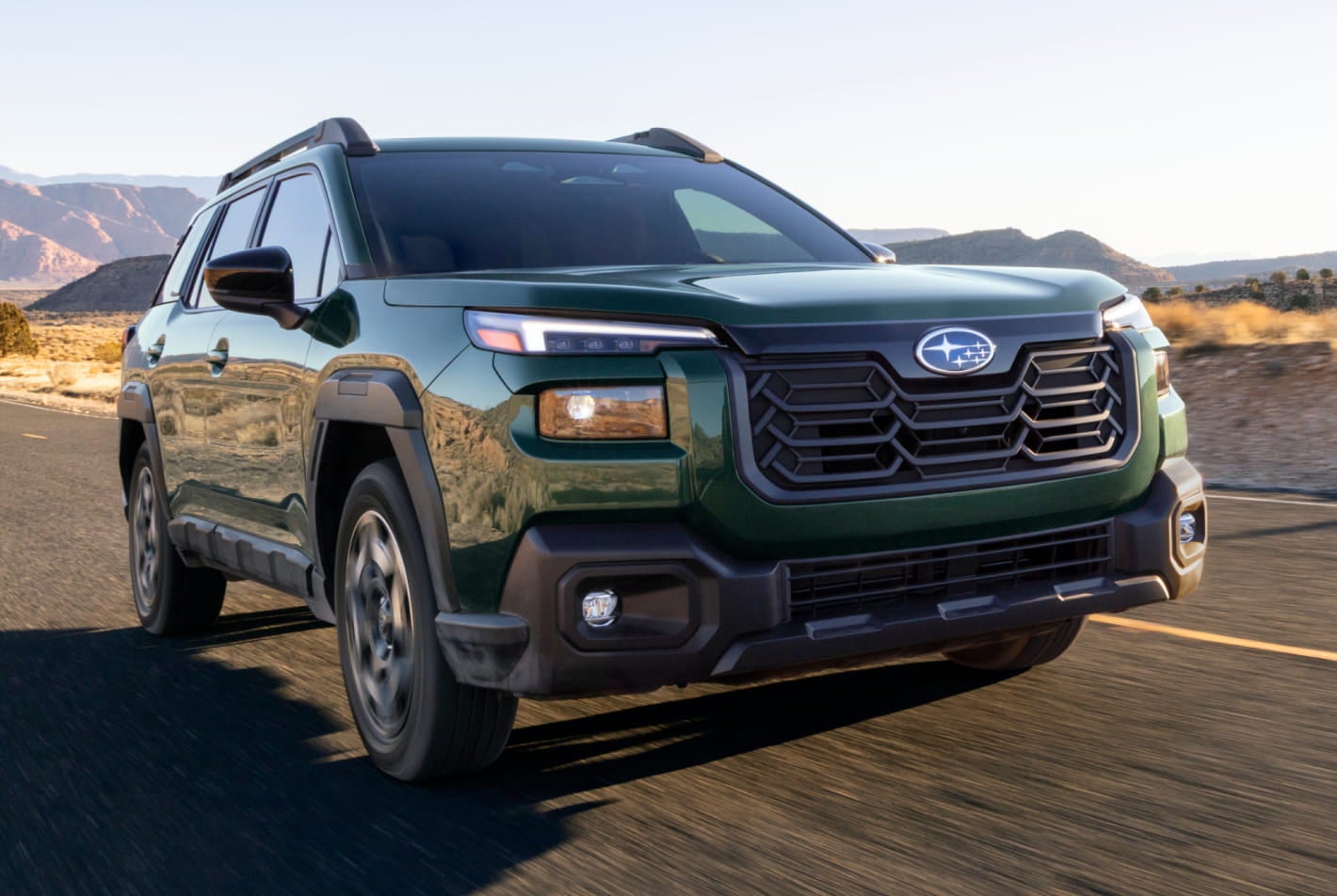 Green Subaru SUV driving on a paved road with an open landscape in the background under a clear sky.