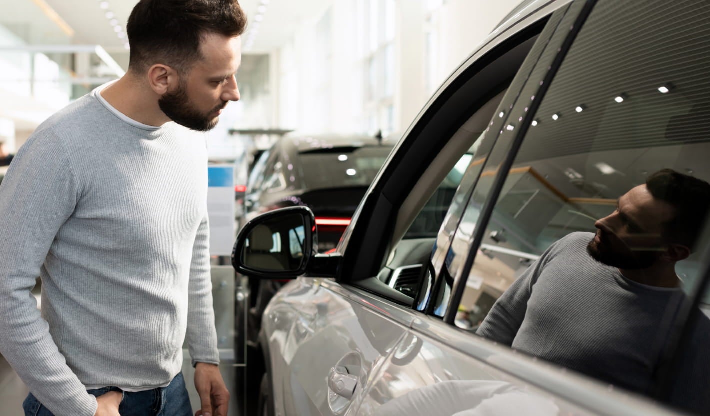 Person inspecting the side window and exterior of a white SUV inside a bright vehicle showroom.