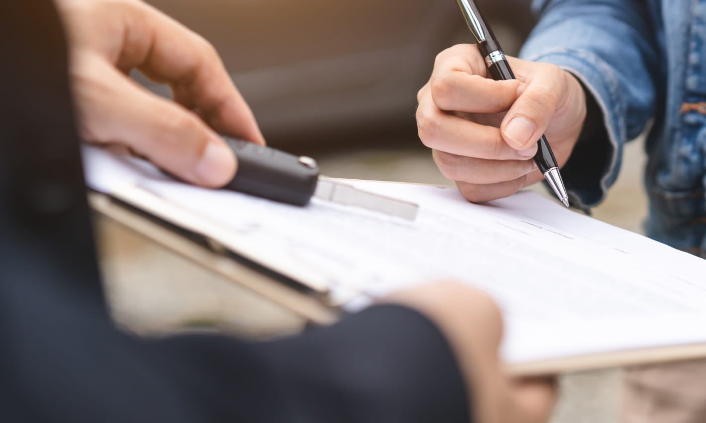 Close-up of a person signing paperwork while another person holds a vehicle key across the table.
