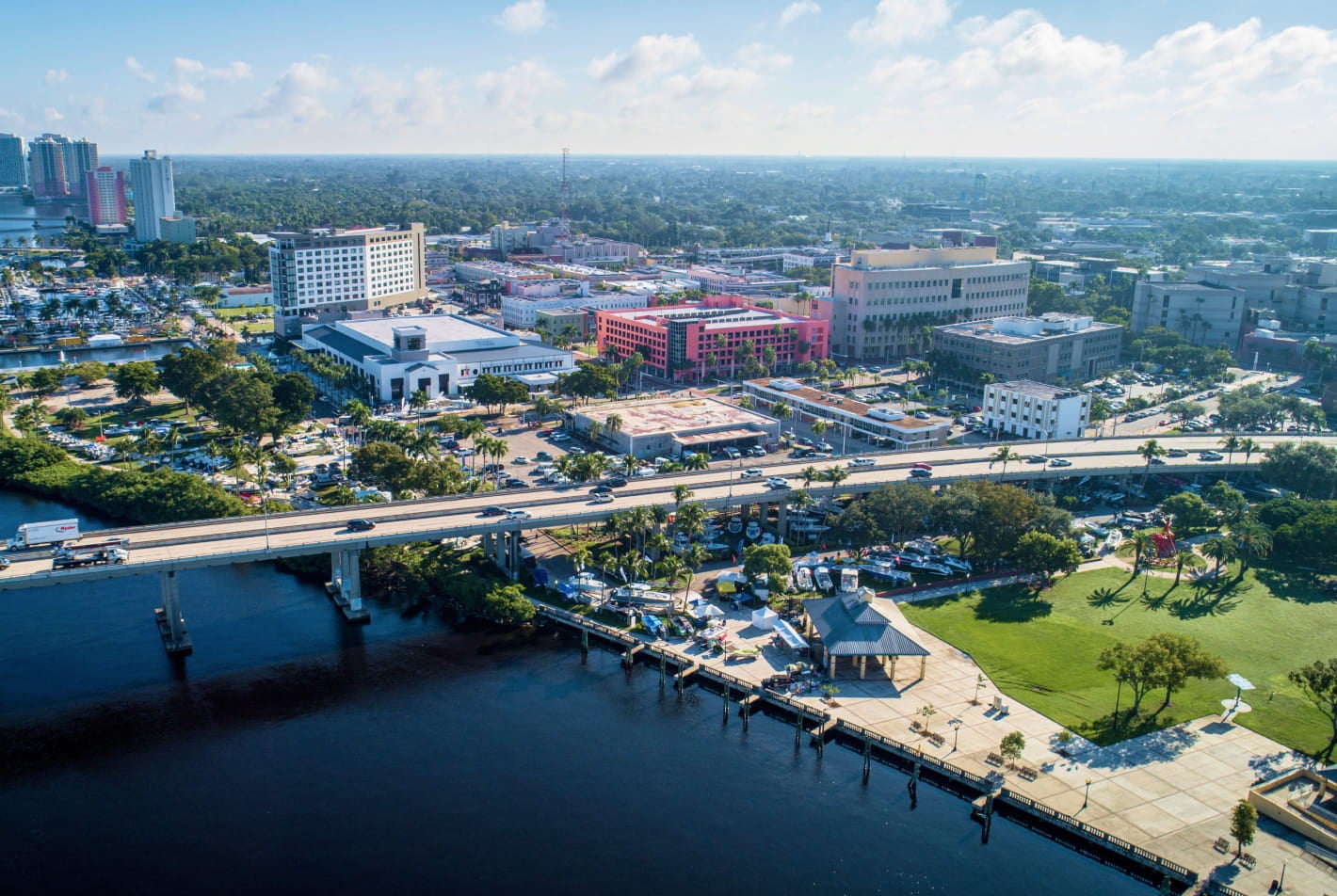 Aerial view of a waterfront cityscape featuring a bridge, downtown buildings, and green spaces under a bright blue sky.