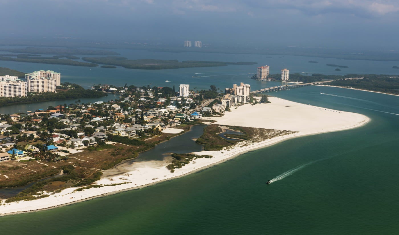 Aerial view of a sandy coastline with residential neighborhoods, waterfront buildings, a bridge, and surrounding turquoise waters.